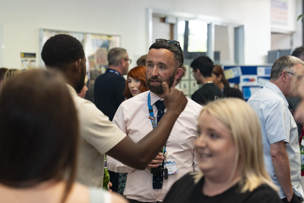 A man in a white shirt and a staff lanyard talks to a man gesturing in a crowded room.