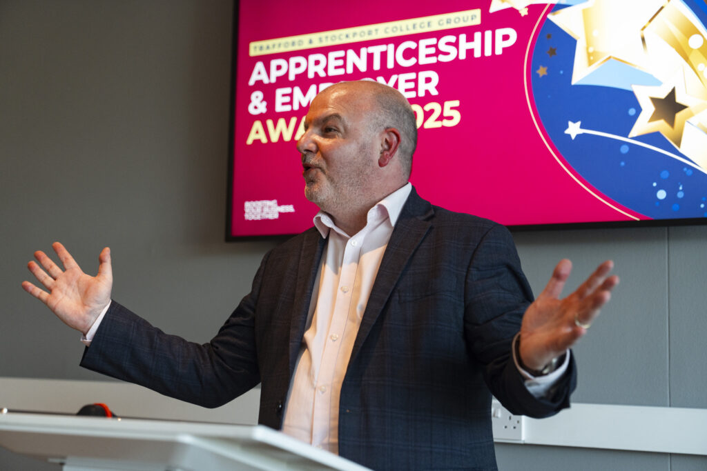 A smiling bald man in a suit gestures with both hands while speaking, with a large screen behind him displaying