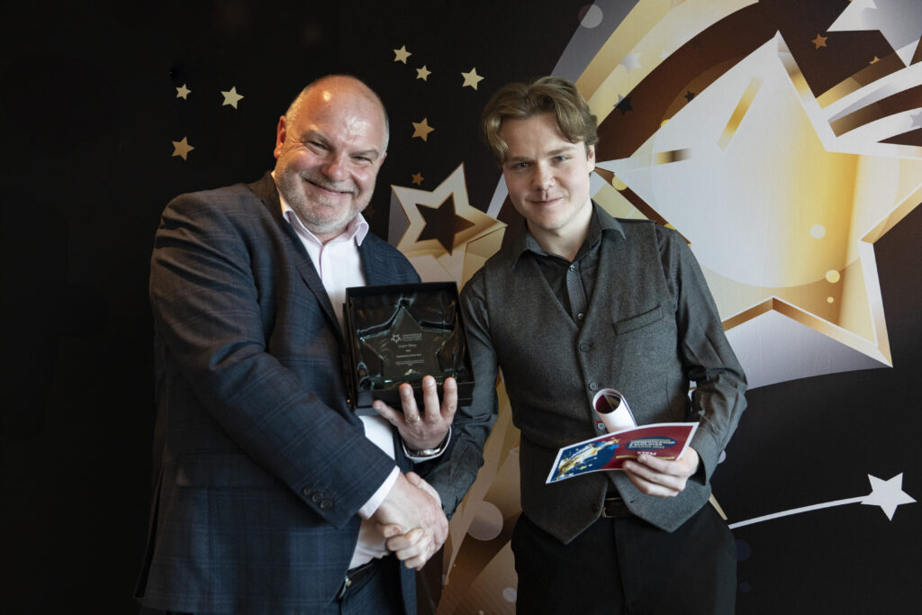 Two smiling men shaking hands, one in a suit holding an award, the other in a shirt holding a certificate, against a star-patterned background.