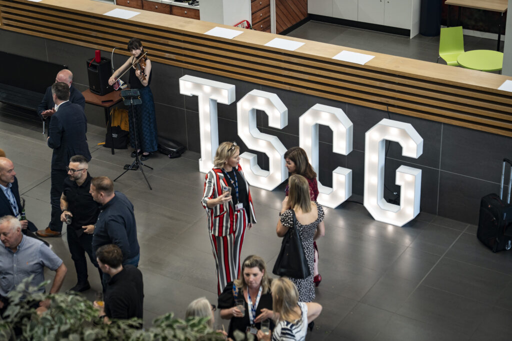 A high-angle view of a gathering with several people mingling. Large illuminated letters spell out "TSCG". In the background, a person plays a violin on a raised platform.