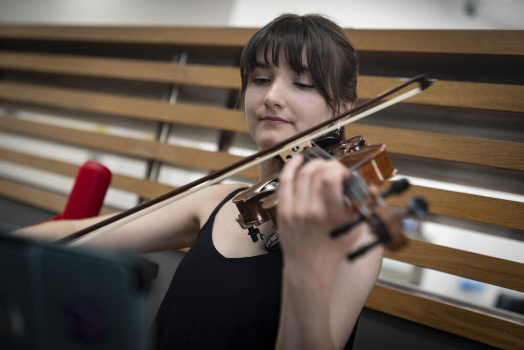 A young woman with dark hair plays a violin with focus, positioned in front of a modern wooden slatted wall.