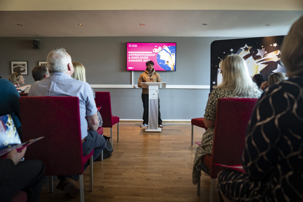 A person stands at a podium addressing an audience seated on red chairs, with a large screen displaying