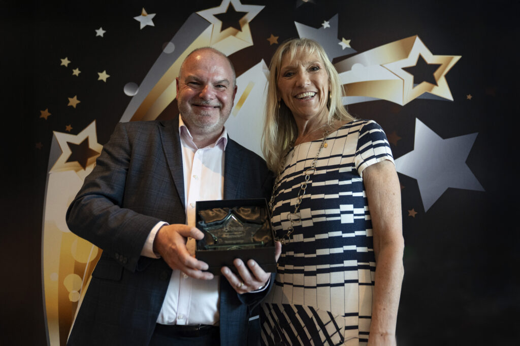 A smiling man in a suit holds a boxed award, standing next to a smiling woman with long blonde hair in a striped dress, against a star-patterned background.