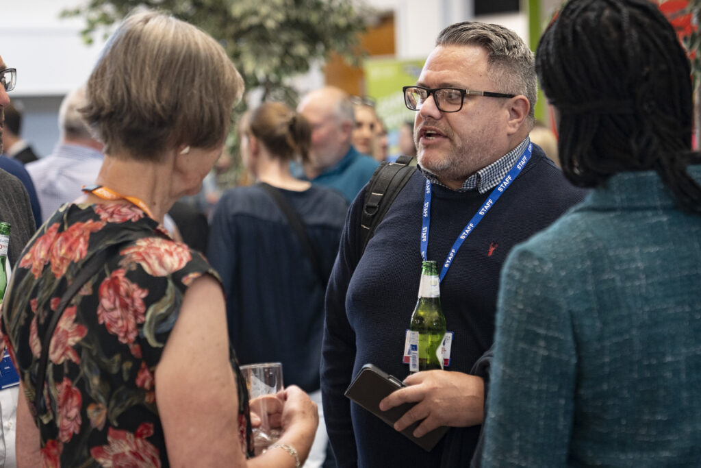 A man in glasses and a staff lanyard talks to a woman in a floral top in a crowded room.